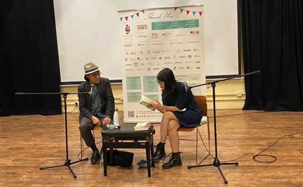 Two people with a microphone each are sitting down and leaning in to talk over a book